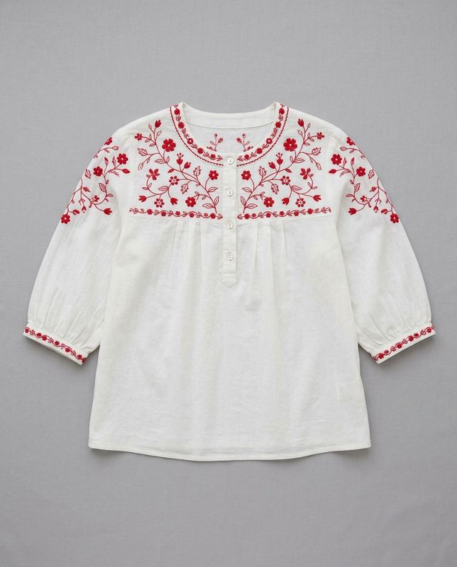 Flat lay of a white blouse with red floral embroidery across the yoke and cuffs