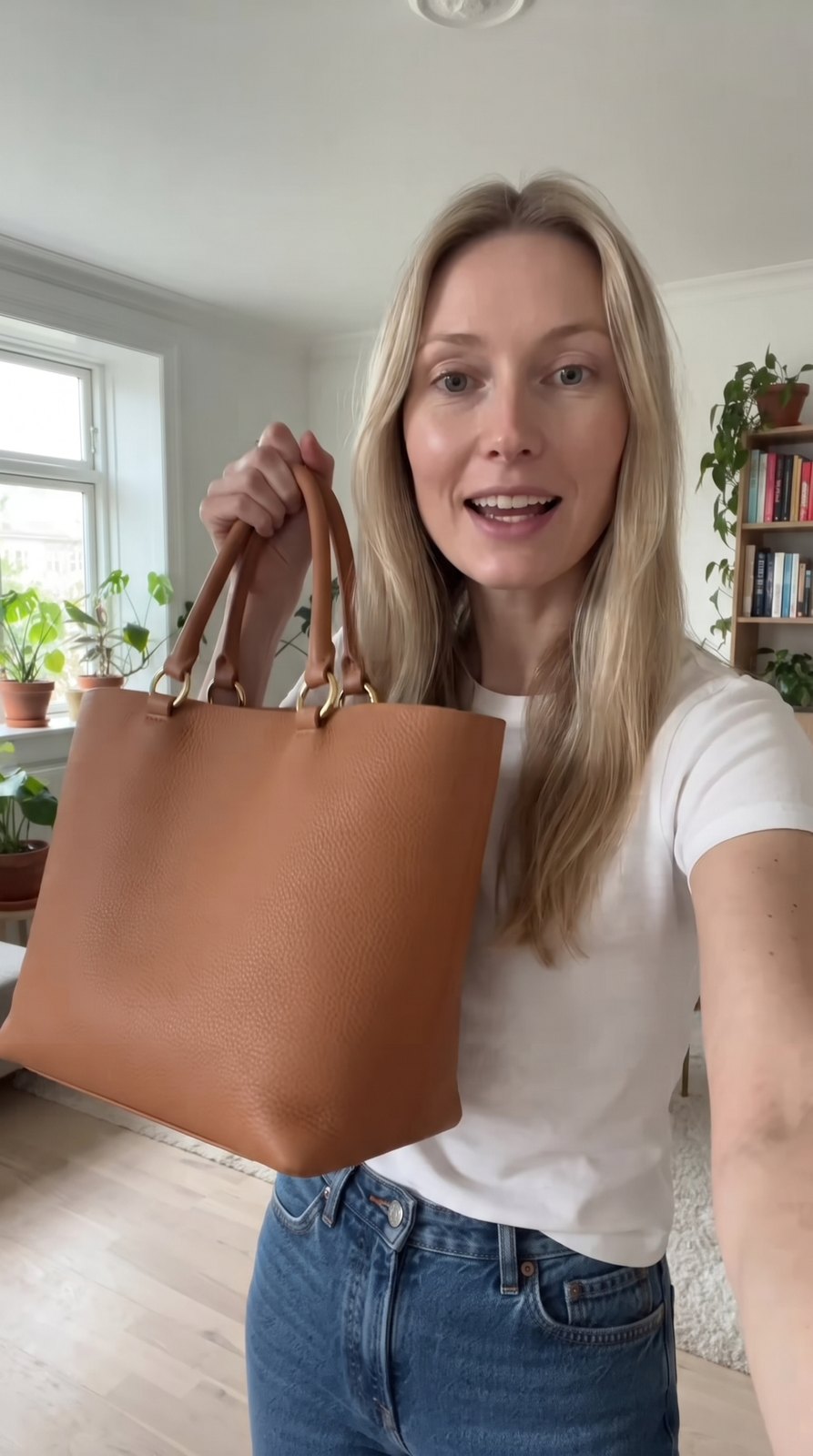 A vertical selfie-style frame of a blonde woman in her apartment holding the tan leather tote up beside her face